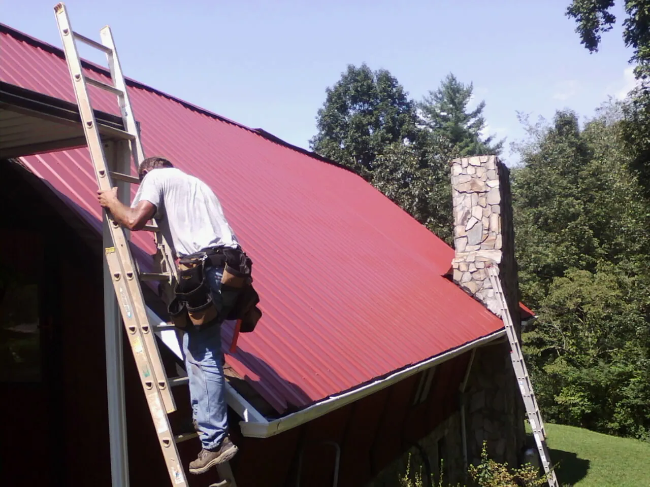 Roofer on a ladder beside a red metal roof and stone chimney — Soffit & Fascia Repair by Roofing Brothers in Sandia Park